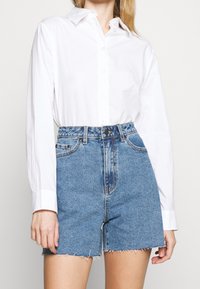 Camisa blanca de manga larga con botones combinada con shorts de mezclilla azul de talle alto, con dobladillo deshilachado y cierre frontal.