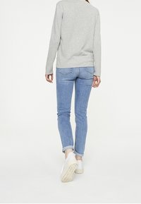 Henkilö kävelee poispäin vaaleanharmaa pitkähihaisessa paidassa, käärityissä sinisissä farkuissa ja valkoisissa lenkkareissa valkoista taustaa vasten.