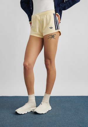 Osoba nosící krémové šortky Adidas s tmavě modrými pruhy a tetováním pavouka na levém stehně, bílé ponožky a bílé slip-on boty.