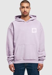 Lilafarbener Kapuzenpullover mit einer Fronttasche und einem weißen quadratischen Logo mit einem Blumenmuster auf der linken Brust. Baumwollmischung, lockere Passform.