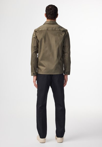 Oliwkowa kurtka o prostym kroju, z dużym kołnierzem i guzikami na mankietach, zestawiona z czarnymi spodniami o prostym fasonie i jasnym obuwiem.