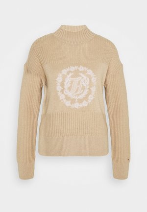 Beżowy dzianinowy sweter z prążkowanym wysokim kołnierzem i rękawami, z białym wyszywanym kwiatowym wieńcem i ozdobnymi inicjałami na piersi.