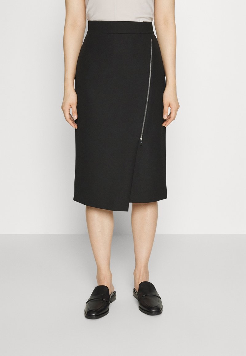 Jupe noire longueur genou avec un détail de fermeture éclair diagonale à l'avant, portée avec des chaussures slip-on noires et un haut clair sur une personne debout.