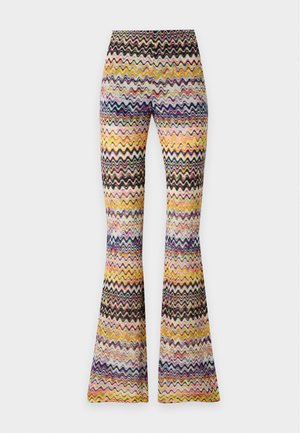Femme aux longs cheveux foncés portant un haut sans manches à motif zigzag coloré et un pantalon évasé assorti, debout avec une main sur la hanche.