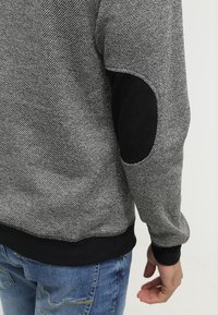 Sudadera gris texturizada con acentos negros, con parches en los codos. Puños y dobladillo negros, combinada con jeans azules.