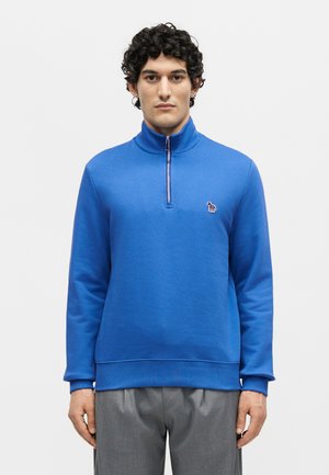 Młody mężczyzna z kręconymi włosami nosi jaskrawo niebieską bluzę z suwakiem do ćwiartki z małym haftowanym logo na lewej piersi, w połączeniu z szarymi spodniami.