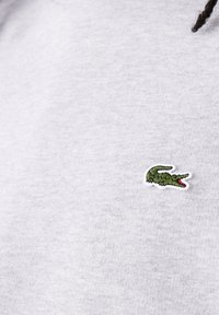Logo crocodile vert brodé sur tissu gris clair, partie d'un vêtement, avec des mèches de cheveux tressés foncés visibles en haut.