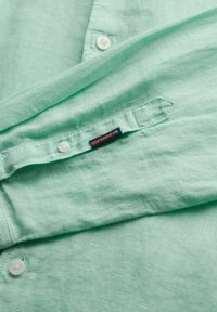 Svetlozelená ľanová košeľa s hladkou textúrou, s bielymi gombíkmi a textilnou nálepkou s nápisom "SUPERDRY" v oranžovej a modrej farbe.
