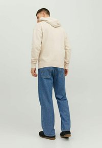 Beżowa bluza z kapturem z kieszenią kangurką, noszona na luźne niebieskie dżinsy. Czarne buty wsuwane o minimalistycznym designie i miękkiej teksturze.