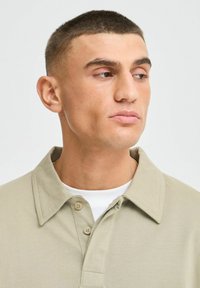 Beige polo shirt met een klassieke kraag, voorzien van een gestructureerde stof, drie knopen, en gedragen over een wit t-shirt.
