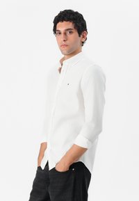 Camisa blanca de mangas largas con cuello abotonado, hecha de un tejido ligero, que presenta un pequeño detalle de logo en el pecho y puños enrollados.