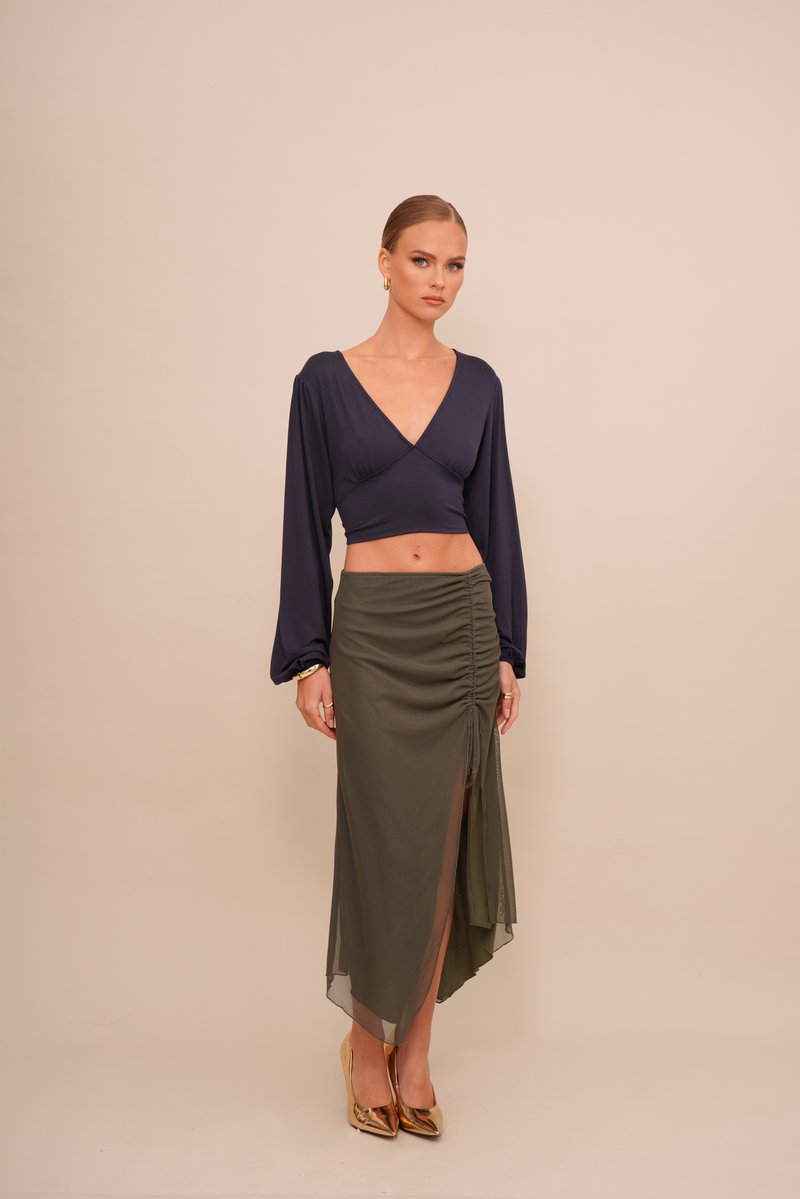 Tengerészeti crop top hosszú ujjakkal és V-nyakkal, zöld olajszínű, ráncolt szoknyával párosítva, amelynek combközépig vágott része és áttetsző rétege van. Arany magassarkú.