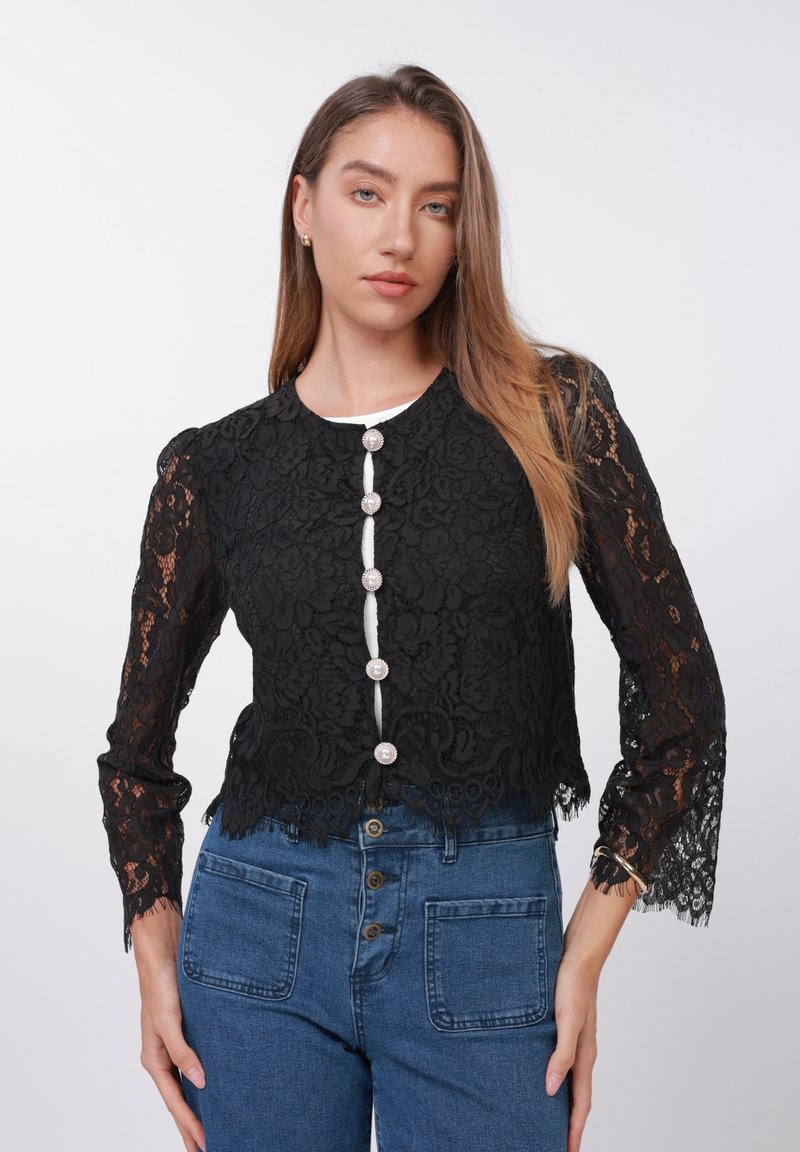 Cardigan negru scurt din dantelă cu model floral, guler rotund, șase nasturi decorativi și mâneci cu franjuri, asortat cu blugi albaștri cu talie înaltă.