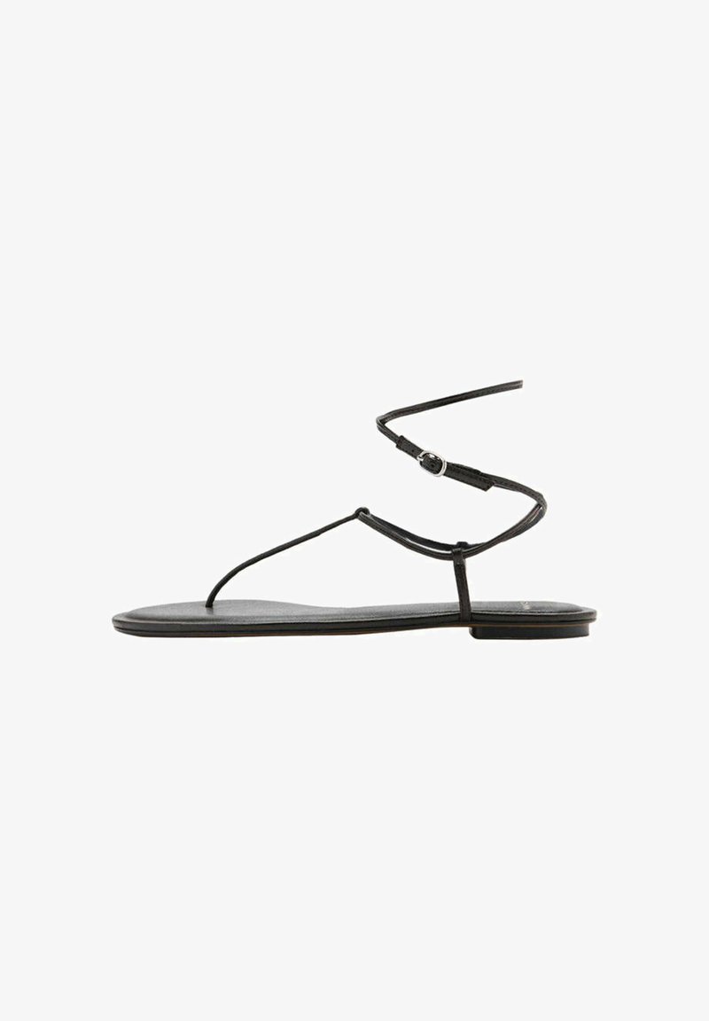 Sandale plate noire avec fines brides et fermeture à boucle autour de la cheville, design minimaliste, vue de côté sur fond blanc.