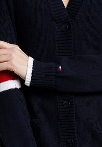 Marineblauwe gebreide cardigan met een diepe V-hals, voorzien van grote knopen en textuurdetails in kabelstructuur. De mouwen hebben rode en witte gestreepte accenten.