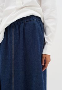Denim krilo z elastičnim pasom, temno modre barve, z svetlim šivom in razširjenim krojem, nosi se z belo dolgimi rokavi.