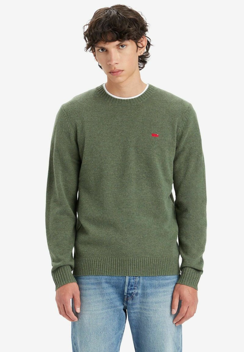 Groene gebreide trui met een ronde hals, geribbelde manchetten en zoom. Heeft een klein rood logo op de borst. Draagt met lichtblauwe jeans.