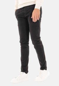 Pantaloni neri slim-fit con una texture liscia, tasche frontali e chiusura con zip, abbinati a sneakers bianche per un look casual.