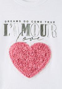 T-shirt bianco in cotone con un cuore rosa testurizzato realizzato in petali di tessuto e testo metallico argento che recita "I SOGNI DIVENTANO REALTÀ L'AMOUR love."
