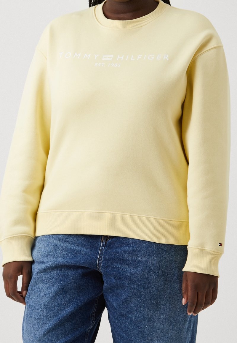 Gelbes Fleece-Sweatshirt mit Rundhalsausschnitt, versehen mit weißem Text "TOMMY HILFIGER EST. 1985" und gerippten Bündchen sowie Saum. Getragen mit blauen Jeans.