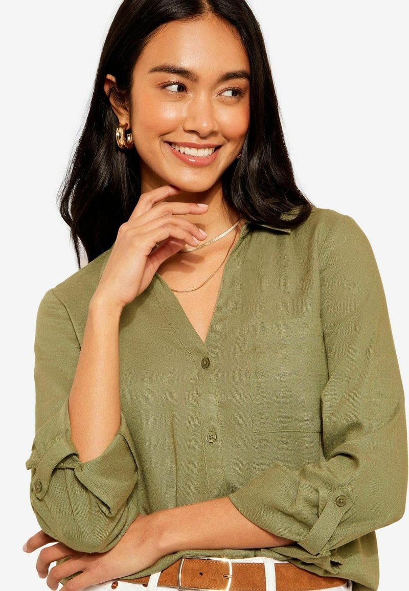 Camicia leggera color verde oliva con bottone, scollatura a V, maniche arrotolate e taschino sul petto. Tessuto liscio e vestibilità comoda.