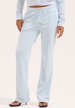 Frau in hellblauen Jogginghosen und passender cropped Jacke, steht mit entspannten Händen, schwarze hohe Sandalen sichtbar.