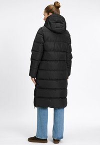 Frau mit hochgestecktem Haar, die einen langen schwarzen Puffer-Mantel mit Kapuze, blaue Jeans und braune Schuhe trägt, steht mit dem Rücken zu einer weißen Hintergrund.