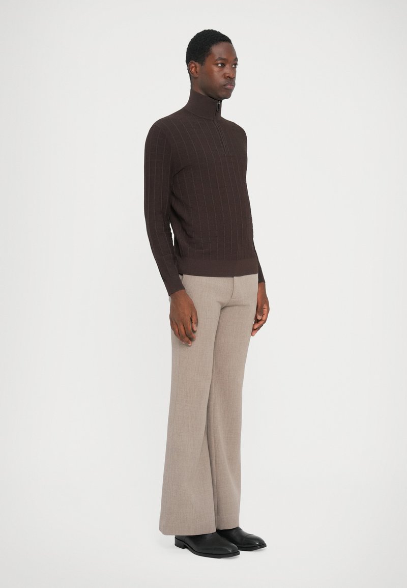Brun, ribbet turtleneck sweater med halvlukning, parret med lysebrune brede bukser og sorte sko. Glat, tekstureret stof.
