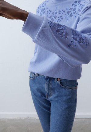 Person trägt einen hellvioletten Pullover mit floralen Spitzen- Ausschnitten an Ärmeln und Brust, kombiniert mit blauen Jeans.