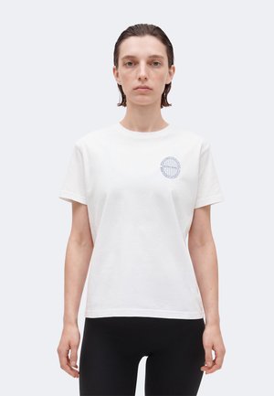 Γυναίκα με κοντά βρεγμένα μαλλιά που φοράει λευκό T-shirt με μικρό κυκλικό λογότυπο στο αριστερό στήθος και μαύρο παντελόνι, στέκεται μπροστά σε απλό φόντο.