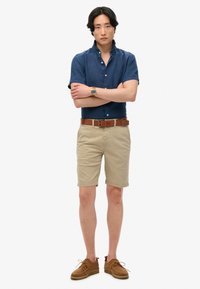 Camicia azzurra a maniche corte con bottoni, pantaloni corti beige, cintura in pelle marrone e scarpe in camoscio marrone. Design semplice con vestibilità rilassata.