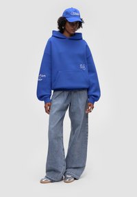 Modrá oversized mikina s přední kapsou, spárovaná se světle modrými širokými džíny a odpovídajícím modrým kšiltem se bílým vyšíváním.