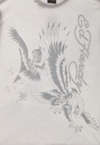Bílý tričko s grafickými designy stříbrných orlů a textem "Ed Hardy." Obsahuje složité vzory peří a detaily.