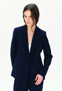 Blazer blu navy con collo a scialle, due tasche frontali, chiusura con un solo bottone e texture del tessuto liscia. Indossato sopra un top in pizzo bianco.