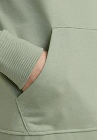 Ljusgrön sweatshirttyg med ribbade muddar och en framficka. Slät textur och sydda detaljer nära fickan.