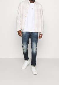 Licht geruite overhemd over een wit T-shirt met "YPS" print, gecombineerd met versleten donkerdenim jeans en witte sneakers.