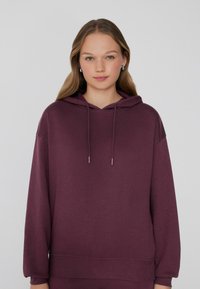 Burgundowa bluza z kapturem z miękkiego materiału, o luźnym kroju, z długimi rękawami i z regulowanym sznurkiem przy dekolcie.