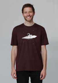 Bordeaux katoenen t-shirt met een witte astronautafbeelding op een vliegerdesign, tegen een sterrenachtergrond. Korte mouwen, ontspannen pasvorm.