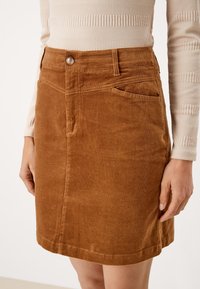 Jupe en velours côtelé marron avec une coupe ajustée, fermeture à boutons sur le devant et poches latérales. Texture lisse et ourlet prenant fin au niveau des cuisses.