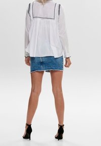 Camicia bianca con bordi e bottoni neri, abbinata a una gonna di jeans blu. Scarpe con tacco nere aggiungono contrasto. Texture dei tessuti lisci visibili.