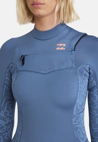 Combinaison de plongée bleue avec une poche zippée, des coutures à plat et des motifs abstraits sur les manches. Présente une texture lisse et un design ajusté.