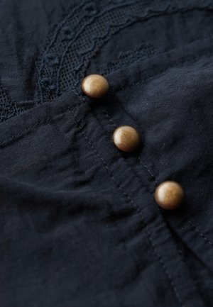 Camicia in tessuto blu navy con dettagli in pizzo decorativo e tre bottoni metallici rotondi in oro. Materiale morbido e testurizzato con cuciture visibili.