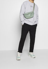 Lichtgrijze trui met handgeschreven logo, zwarte broek en groene schoudertas, gecombineerd met witte en mintkleurige sneakers.