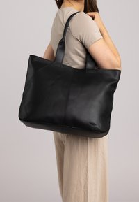 Zwarte leren tote bag met gebogen naden en stevige handvaten. Beschikt over een gladde textuur en een subtiel logo dat aan de onderkant is geperst.