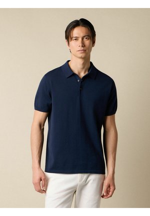 Man in een marineblauw korte mouwen poloshirt en witte broek, staand tegen een effen beige achtergrond.