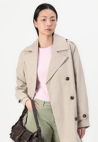 Beige trenchcoat med dobbeltraders front, stor krage og mørke knapper. Brukt over en rosa skjorte og grønne cord-bukser, med en brun veske.