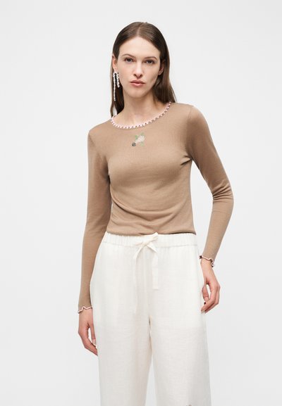 Kvinde iført beige langærmet top med lyserød kant ved halsudskæringen og hvide bukser med snøre, poserer foran en ensfarvet hvid baggrund.