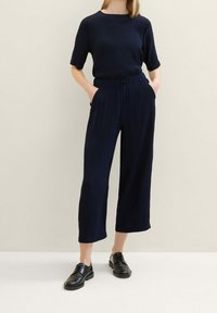 Žena oblečená v námořnickém strukturovaném topu a odpovídajících širokých zkrácených kalhotách, s černými šněrovacími botami, stojící s rukama v kapsách.