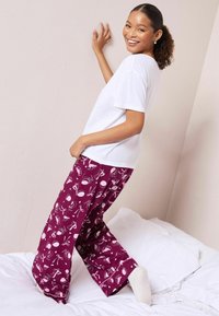Hvid T-shirt med korte ærmer, parret med maroon pyjamasbukser, der har et legende mønster af forskellige genstande. Modell står på en seng.