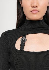 Črn rebrast turtleneck s izrezom in dekorativno srebrno zaponko. Tkanina se zdi teksturirana in prilegajoča.